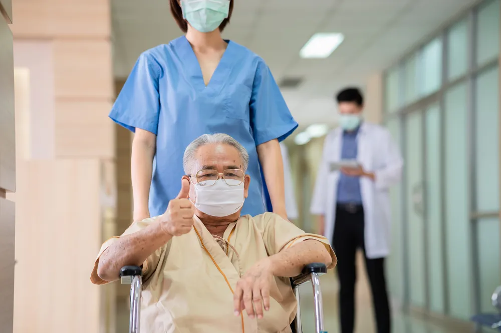 Portrait of senior Asian man wear protective face mask sitting on wheelchair and recovering from illness showing thumbs up