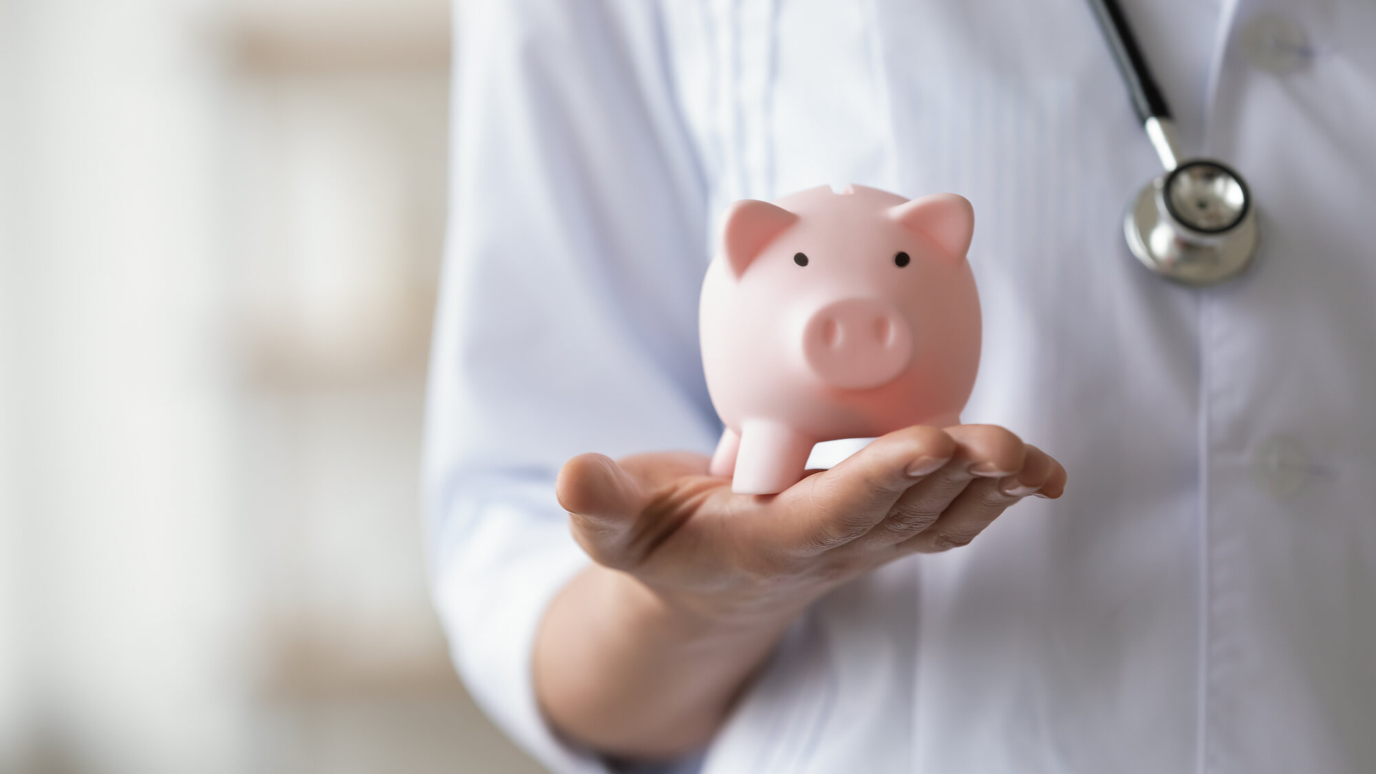 A doctor wearing a stethoscope around their neck, holding a piggy bank in one hand.