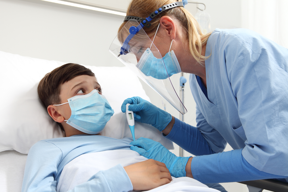nurse with thermometer measures fever on patient child in hospital bed, wearing protective visor and surgical mask, corona virus covid 19 protection concept
