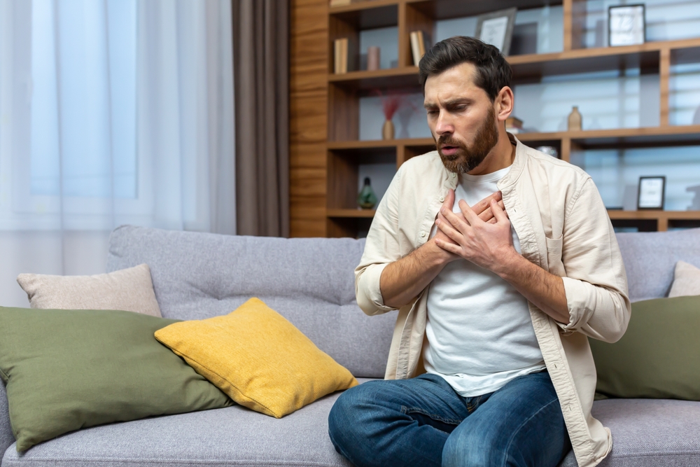 A young man is sitting on the sofa at home and holding his chest