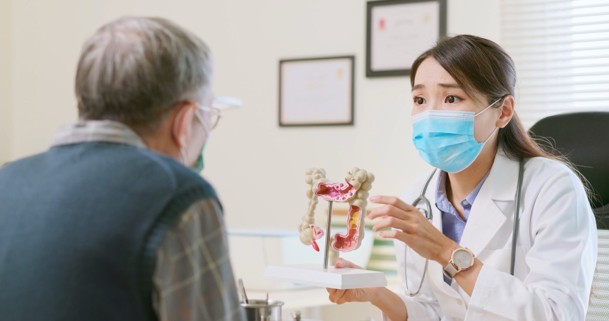 asian female doctor wearing face mask is showing a model of the large intestine and explaining to elder senior man patient in hospital