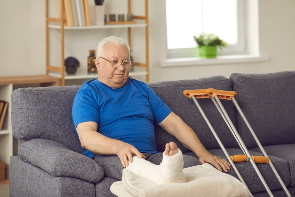 Rehabilitation after domestic or car accident injury: Senior male stays at home, waiting for bone fracture to heal. Old man with broken leg in plaster cast sitting on sofa with crutches in living-room 